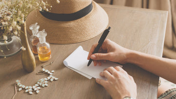 Woman sitting at a vanity table writing gentle confidence affirmations in a notebook, reflecting softly and grounding herself — Talk2Tessa.