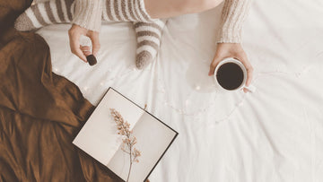 Soft image of a woman sitting on a bed with tea, chocolate and a notebook — a calm moment of January journaling, reflection and gentle self-care.
