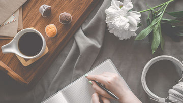 A woman sitting on a bed journaling with tea and small cakes beside her, capturing a soft, calming moment that reflects mental health, self-care and gentle reflection.