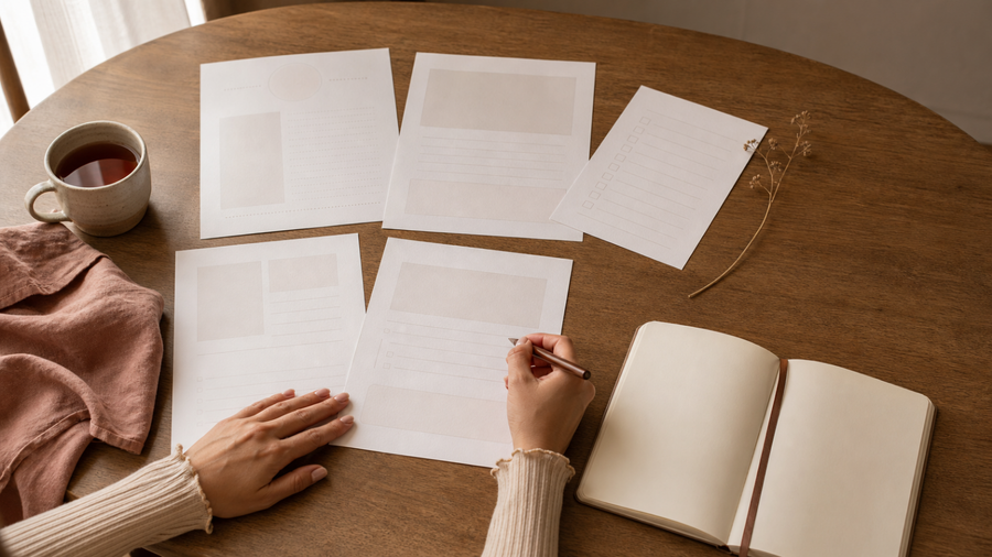Five blank worksheet pages, an open journal, tea, dusty-rose linen, and a woman's hands holding a pen on a warm wooden table.
