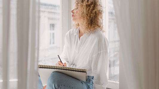 Calm woman writing kindness quotes in her journal by a sunlit window, gentle self-care and reflection time – Talk2Tessa psychologist-designed burnout and self-compassion support.