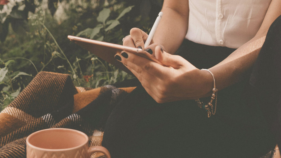 Woman writing gentle affirmations in a quiet outdoor field, capturing a moment of calm self-reflection and emotional softness — Talk2Tessa.