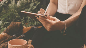 Woman writing gentle affirmations in a quiet outdoor field, capturing a moment of calm self-reflection and emotional softness — Talk2Tessa.